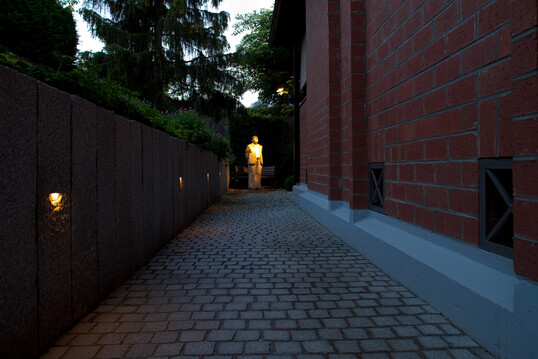 Ein Seitenzugang zum Garten wird durch Stufenleuchten, die in Granitpalisaden eingesetzt wurden, angenehm beleuchtet. Eine originelle Skulptur wird durch eine geschickte Beleuchtung zum Zielpunkt des Weges.